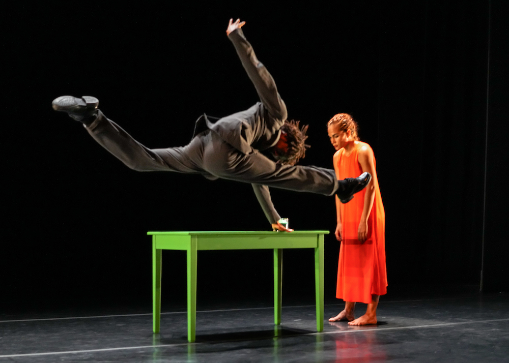man in black flies across the green table in a split jump as a woman in bright orange standing across from him leans foward expressionless 